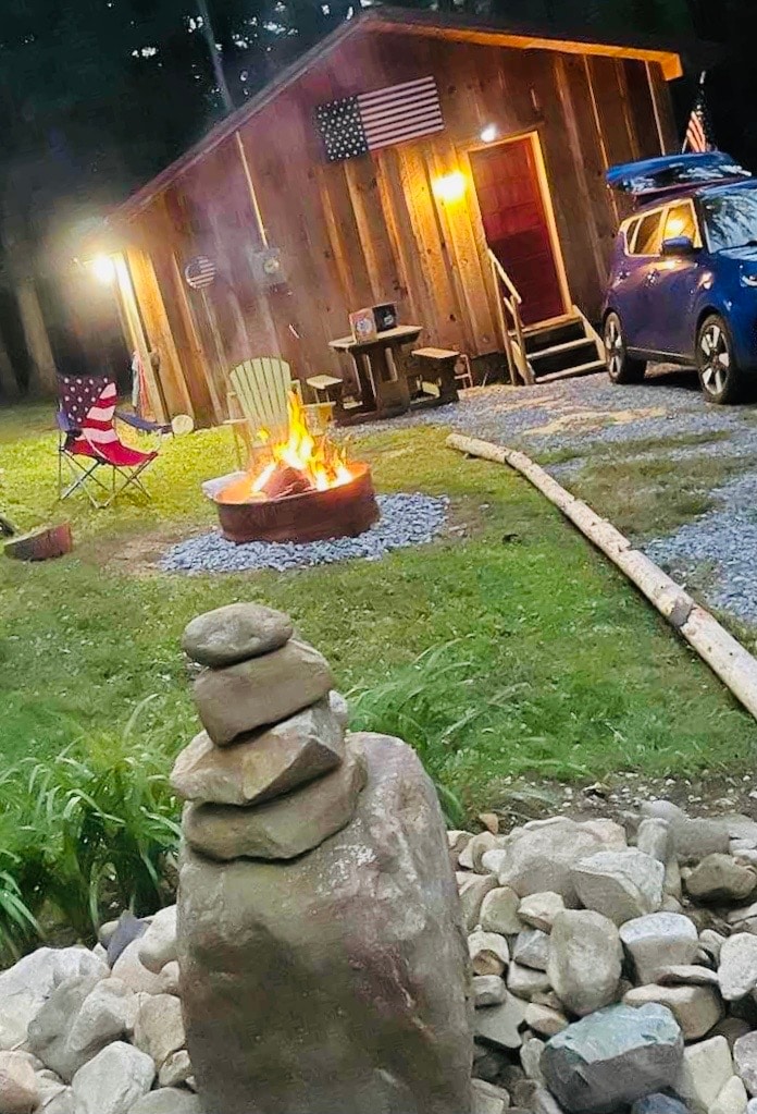 A rustic cabin is illuminated by warm lights, with flags displayed prominently. A fire pit surrounded by stones burns in the foreground, while two chairs and a small table invite relaxation. A vehicle is parked nearby on a gravel path lined with greenery.
