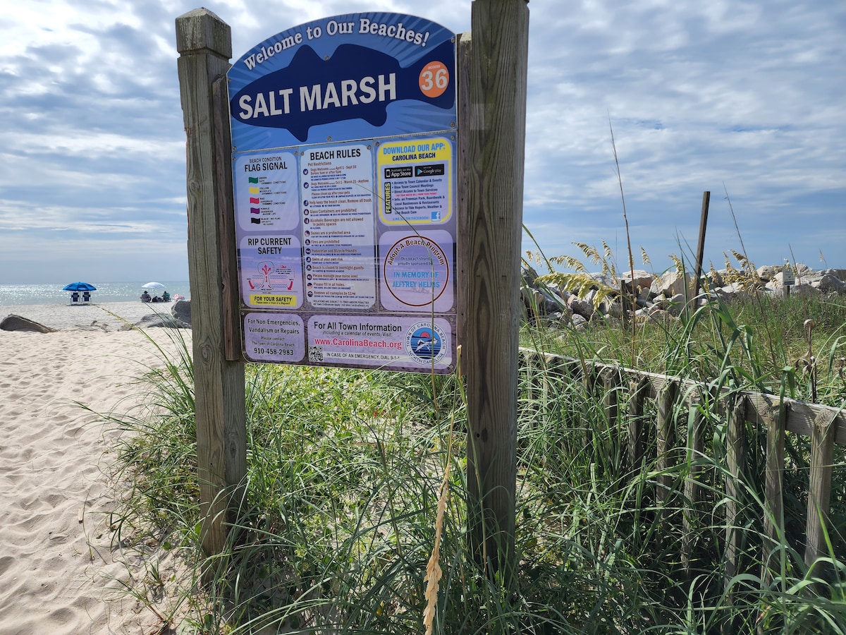 A wooden sign marks the entrance to Salt Marsh, displaying beach rules and information for visitors. Tall grasses frame the sign, and sandy paths lead towards the beach in the background, with a glimpse of the ocean and beach umbrellas visible.