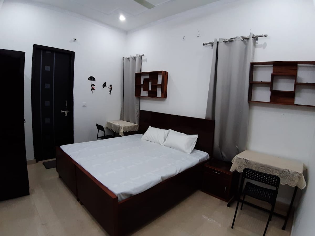 A bright room features a spacious bed with light bedding, complemented by wooden bedside tables and a dresser. Two tables with chairs are positioned in the room. Simple wall shelves provide additional storage, and soft, neutral curtains frame the windows.