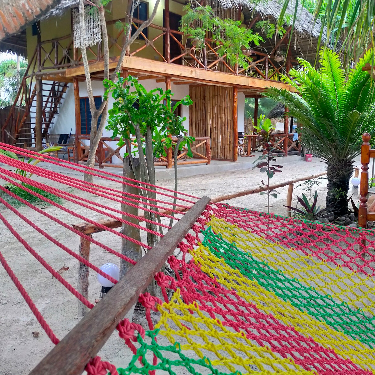 A colorful hammock is draped between wooden posts, offering a place to relax in the shared garden area. The vibrant red, yellow, and green netting contrasts with the sandy ground. In the background, traditional Swahili-style buildings are visible, surrounded by lush greenery and palm trees.