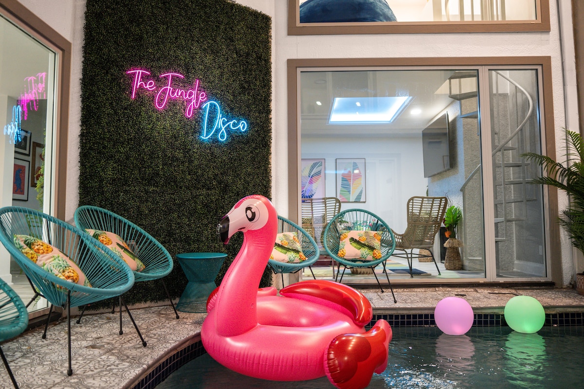 The pool area features a vibrant pink flamingo float, with colorful orbs floating on the water. Comfortable chairs surround the pool, adorned with decorative cushions. A neon sign reading 'The Jungle Disco' adds a playful touch, while large glass doors provide views into the indoor space.