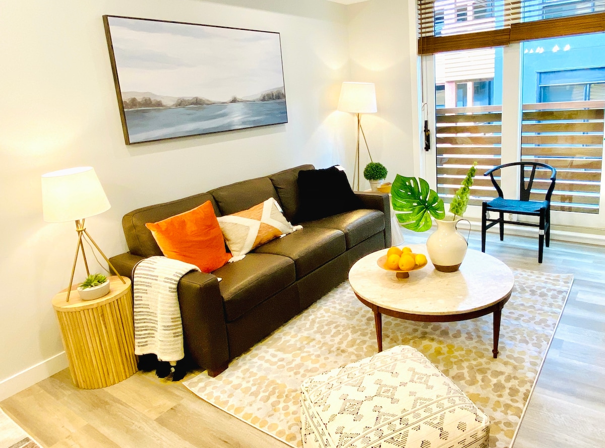 A modern living area is furnished with a dark leather sofa, accented by patterned throw pillows and a soft blanket. A circular coffee table rests in front, surrounded by a plush area rug. Natural light enters through large glass doors leading to a balcony.