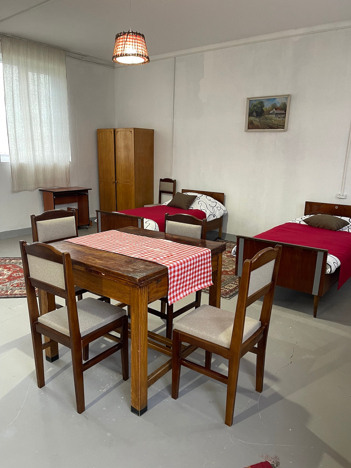 Two single beds with patterned blankets are positioned against the wall, complemented by a dining area featuring a wooden table and four chairs. A cabinet stands nearby, while a landscape painting adorns the wall. Natural light filters through sheer curtains, enhancing the room's spacious feel.