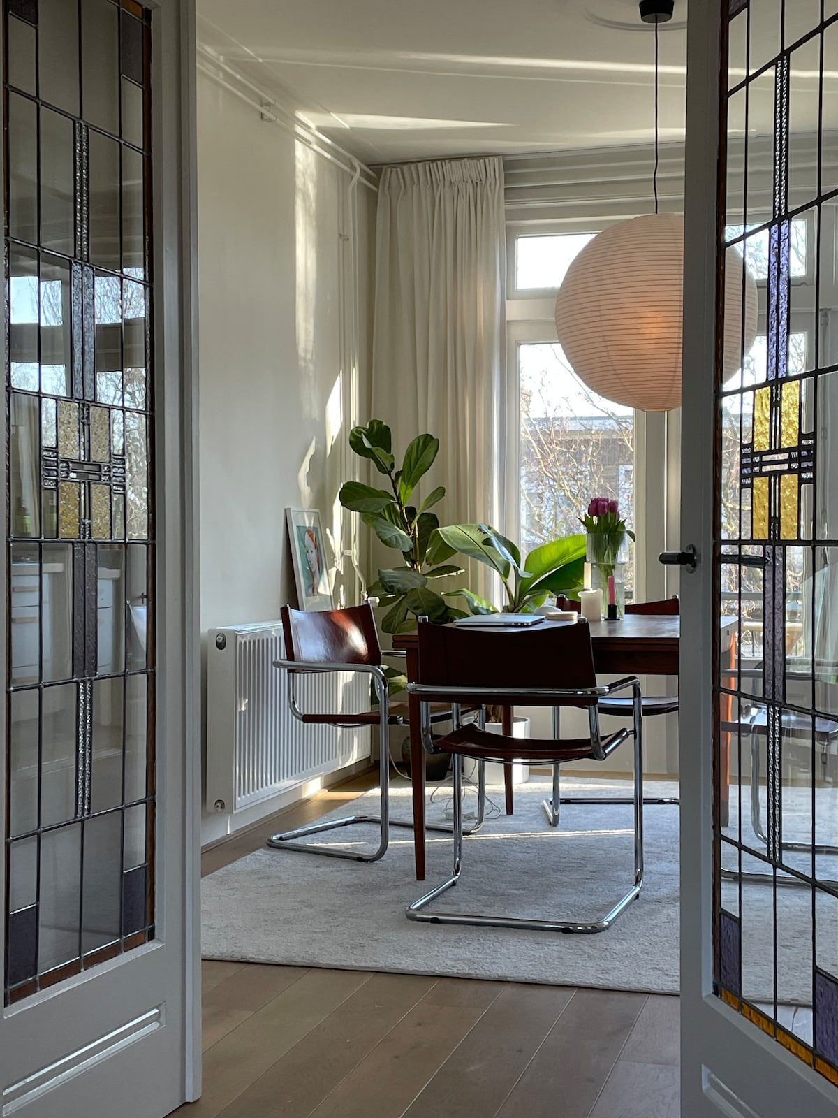 The dining area is illuminated by natural light from large windows, featuring a round pendant light overhead. A neatly set table is flanked by two modern chairs, with greenery adding a touch of nature to the space. A warm wooden floor enhances the inviting ambiance.