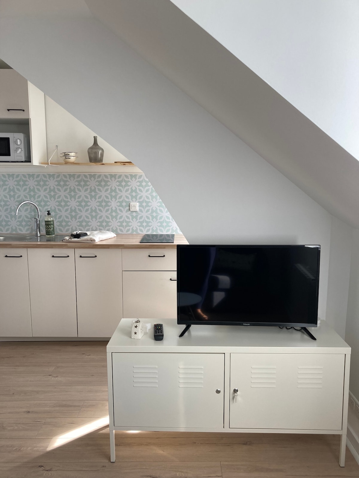 An open living area features a modern television atop a sleek white media console. A kitchenette with minimalistic cabinetry and patterned backsplash is visible in the background. Natural light illuminates the space, highlighting the wooden flooring and angled ceiling.