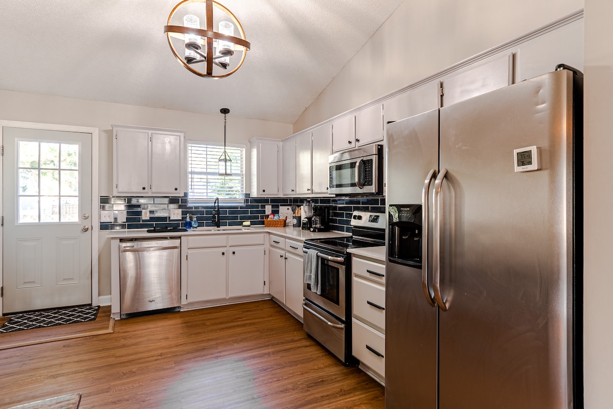 The kitchen features modern stainless steel appliances, including a double-door refrigerator and a dishwasher. White cabinets and dark countertops provide contrast. Natural light enters through a window above the sink, while wood flooring adds warmth to the space. A circular light fixture hangs from the ceiling.