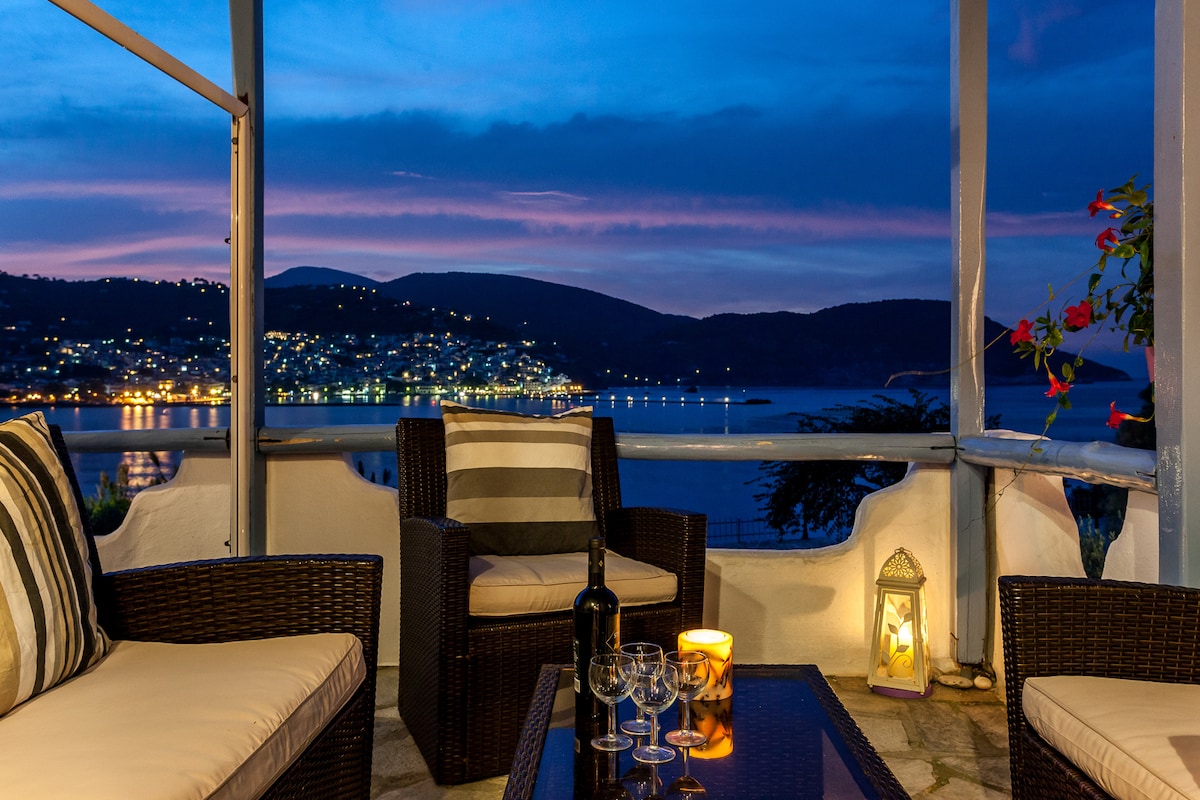 An outdoor seating area is featured, consisting of dark wicker chairs with striped cushions and a glass-topped table. A bottle of wine and glasses are placed on the table. The sea view is illuminated by soft evening light, with the village lights twinkling in the distance.