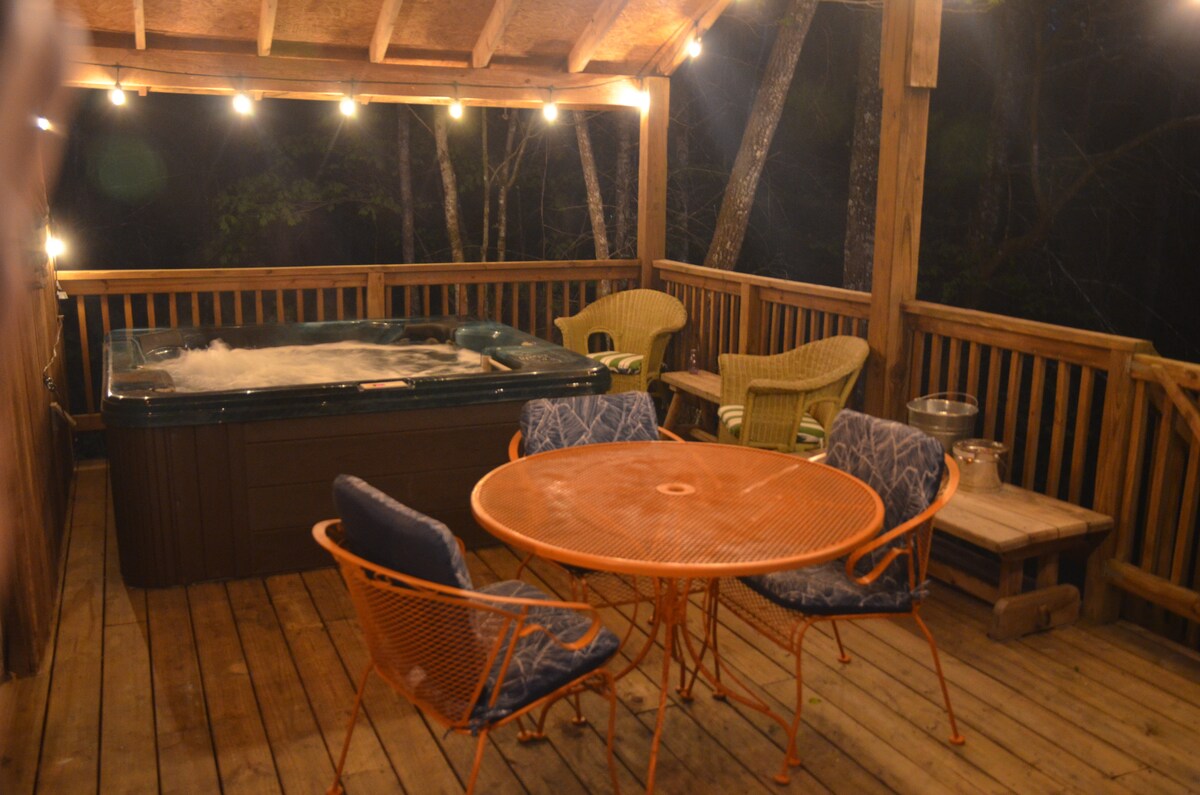A spacious deck area features a hot tub alongside a round dining table with orange metal chairs. Two green woven chairs are positioned nearby, creating a relaxing outdoor space surrounded by trees, illuminated by warm string lights overhead.