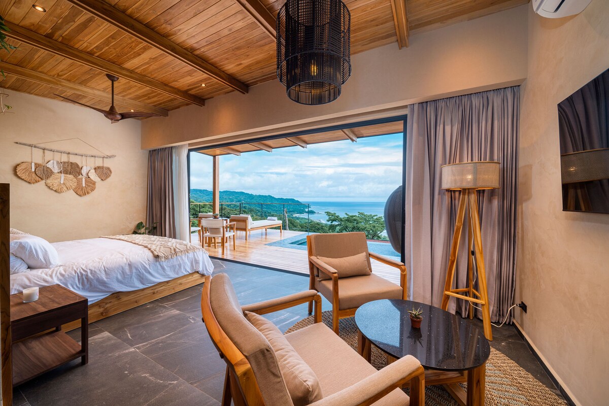 A spacious bedroom features a large bed with crisp white linens, complemented by natural wooden accents. Large glass doors offer scenic ocean views, while a seating area includes two chairs and a round table. Soft lighting and neutral tones create a serene environment.