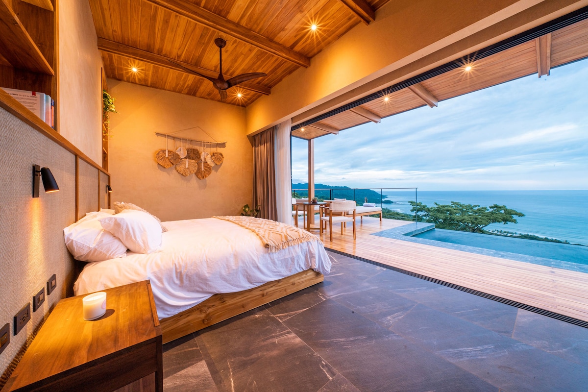 A spacious bedroom features a large bed with white linens and wooden accents. Floor-to-ceiling glass doors open to a private outdoor area, offering expansive views of the ocean. Minimalistic shelves and soft lighting create a serene ambiance, with natural materials enhancing the design.