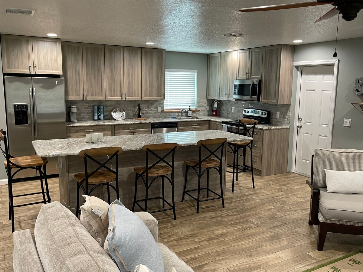 The kitchen features a spacious island with seating for four and modern appliances, including a refrigerator and microwave. The light-colored cabinets and granite countertops offer a contemporary feel. Natural light enters through a window, enhancing the inviting atmosphere of this open layout area.
