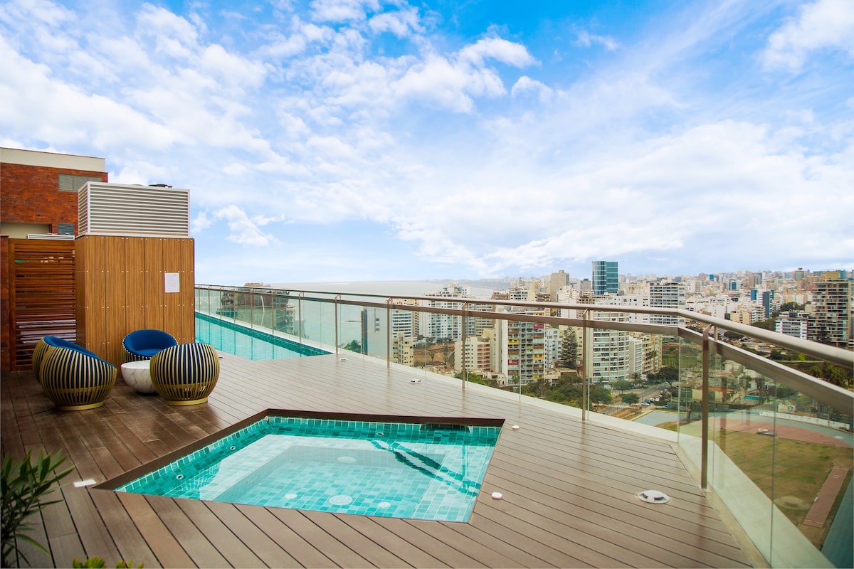 A rooftop area features a small, inviting pool surrounded by wooden decking and glass railings. Two modern chairs are positioned nearby, offering views of the expansive cityscape and sky. The scene captures a blend of urban architecture and open air.