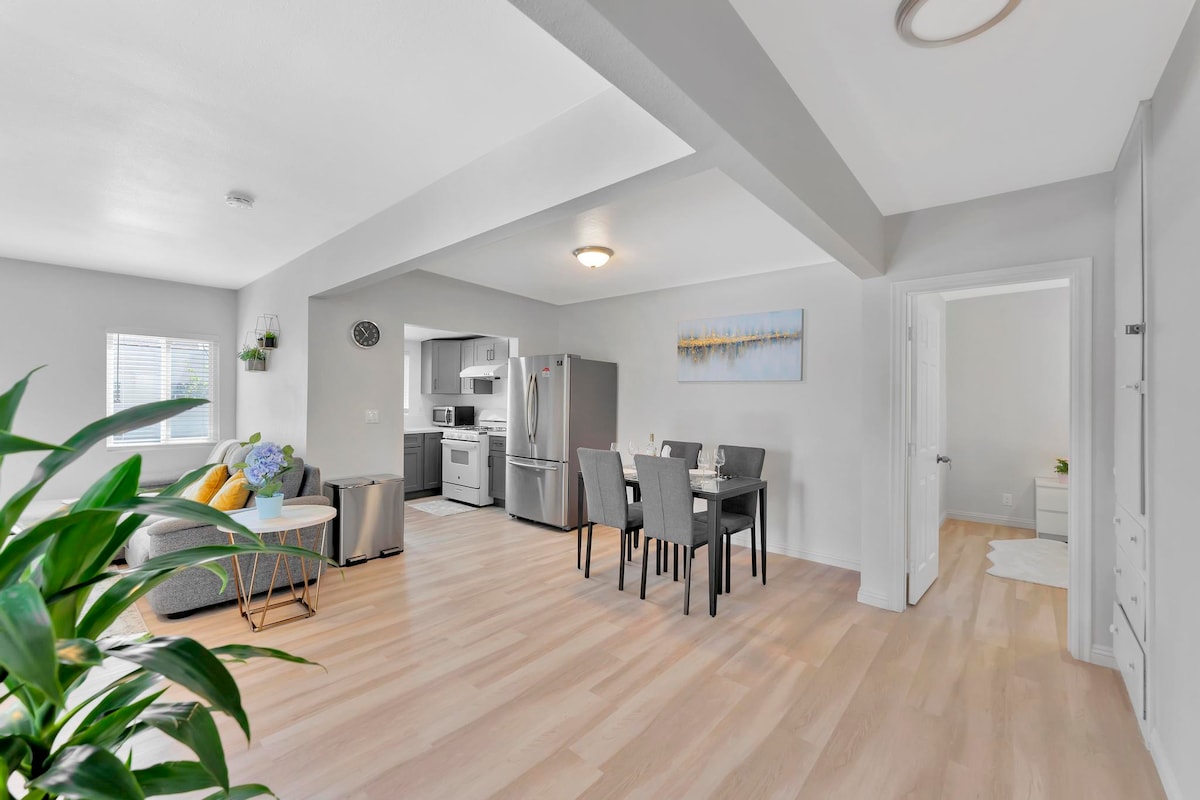 An open living space features light wood flooring and a comfortable seating area with a large grey couch. A dining table for four is positioned nearby, with a modern kitchen visible in the background. Soft natural light filters through the windows.