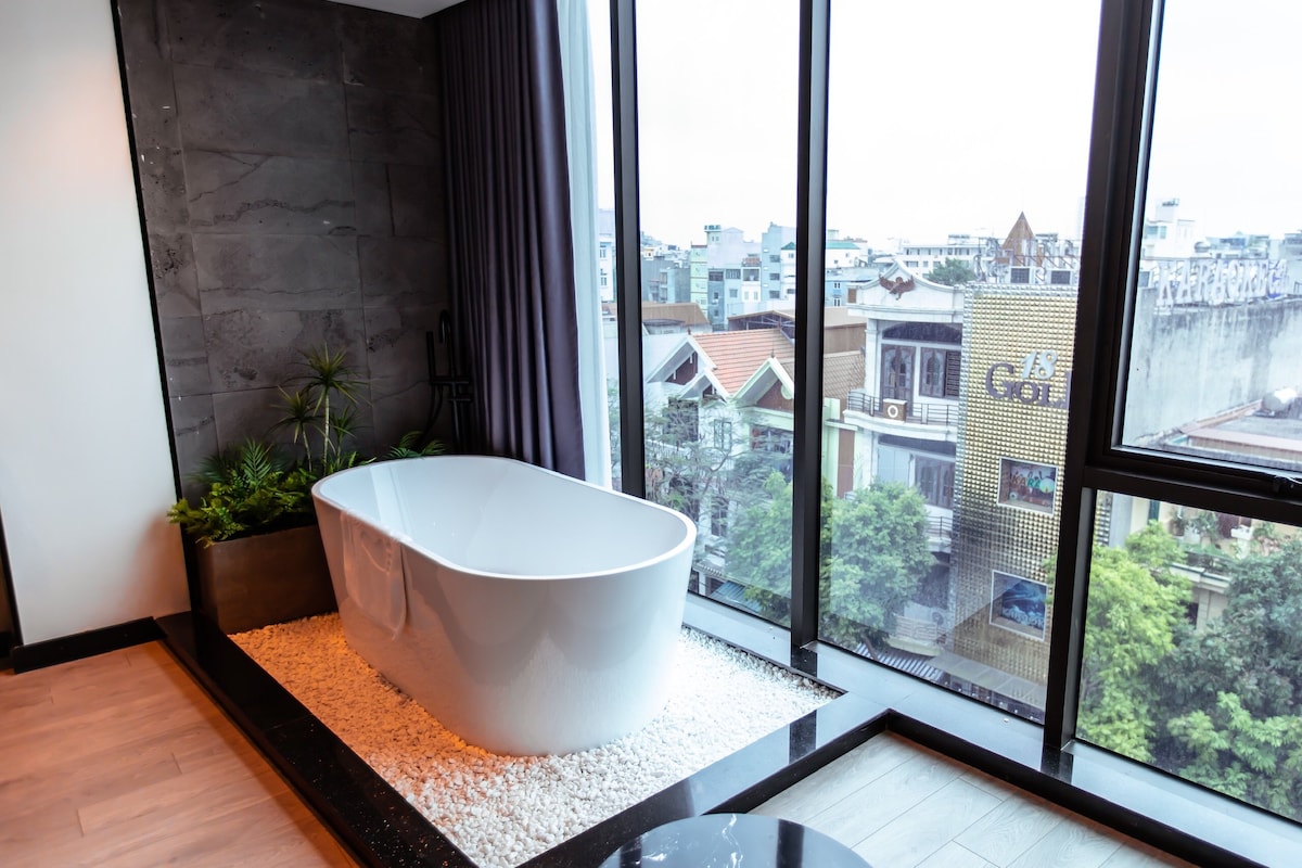 A modern freestanding bathtub is placed on a bed of smooth stones, surrounded by small plants. Large windows offer a view of the city, allowing natural light to fill the space, with dark gray walls providing a subtle contrast.