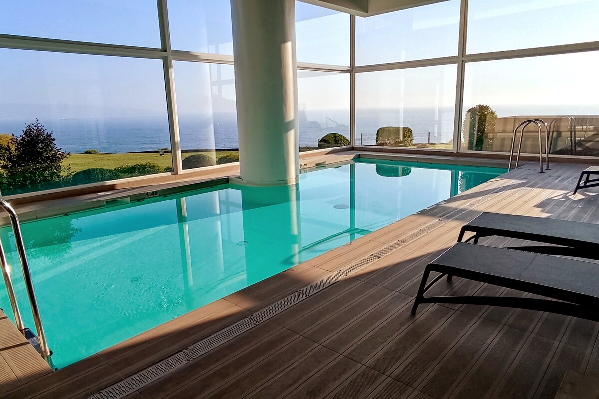 An indoor heated pool is seen with clear turquoise water, surrounded by large glass windows that frame a scenic ocean view. Sunlight casts reflections across the pool's surface, and lounge chairs are positioned nearby on the wooden deck.