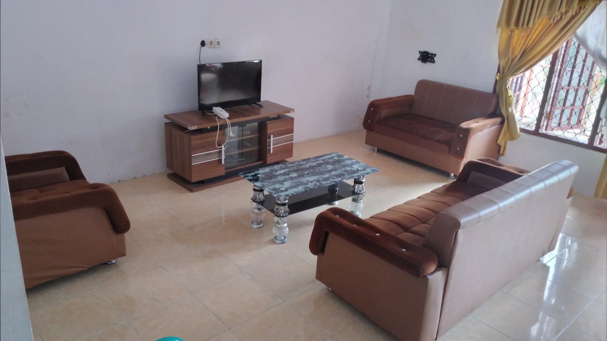 The living area features a television mounted on the wall across from a low glass coffee table, surrounded by two brown leather sofas and a matching loveseat. Natural light enters through a window adorned with elegant curtains.