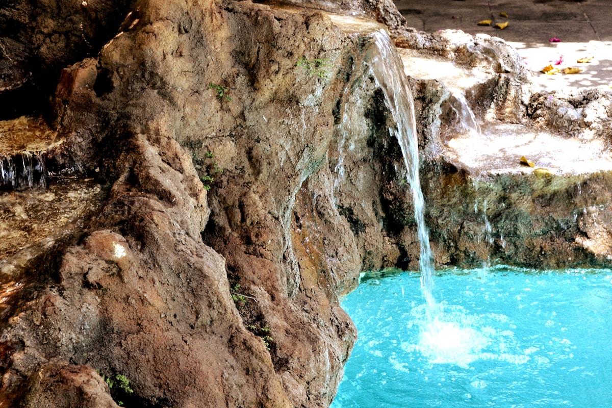 The image features a natural stone structure with a small waterfall cascading into a clear turquoise pool. The rocks exhibit a rugged texture, while the water creates gentle ripples as it flows into the pool, offering a refreshing and tranquil ambiance.