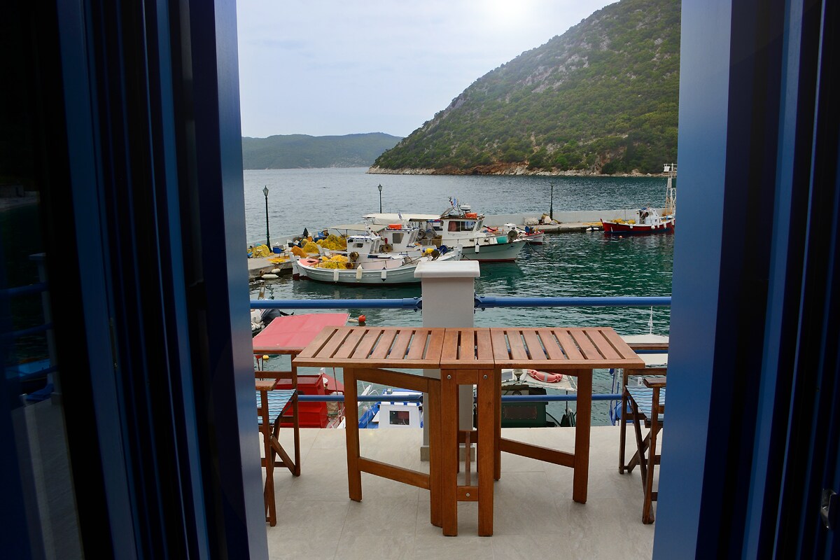 A view of the serene waters of Polis Bay is presented from a balcony. A wooden dining table with matching chairs overlooks a harbor filled with colorful boats, framed by a scenic hillside. Bright sunlight streams gently onto the space.