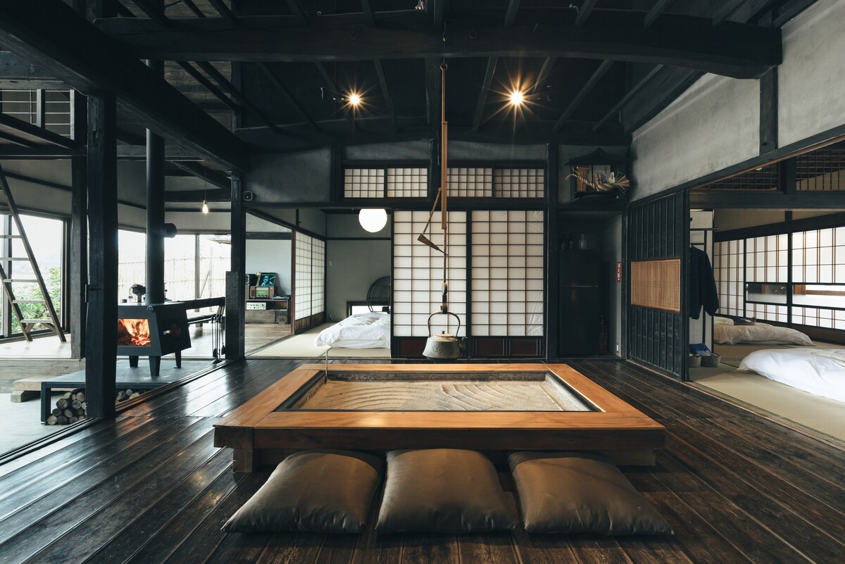 The interior features traditional elements, including exposed wooden beams and a central irori hearth surrounded by plush cushions. Sliding shoji screens create defined spaces, with warm lighting enhancing the room's ambiance. A kitchen area is visible, highlighting the rustic charm of the renovated space.