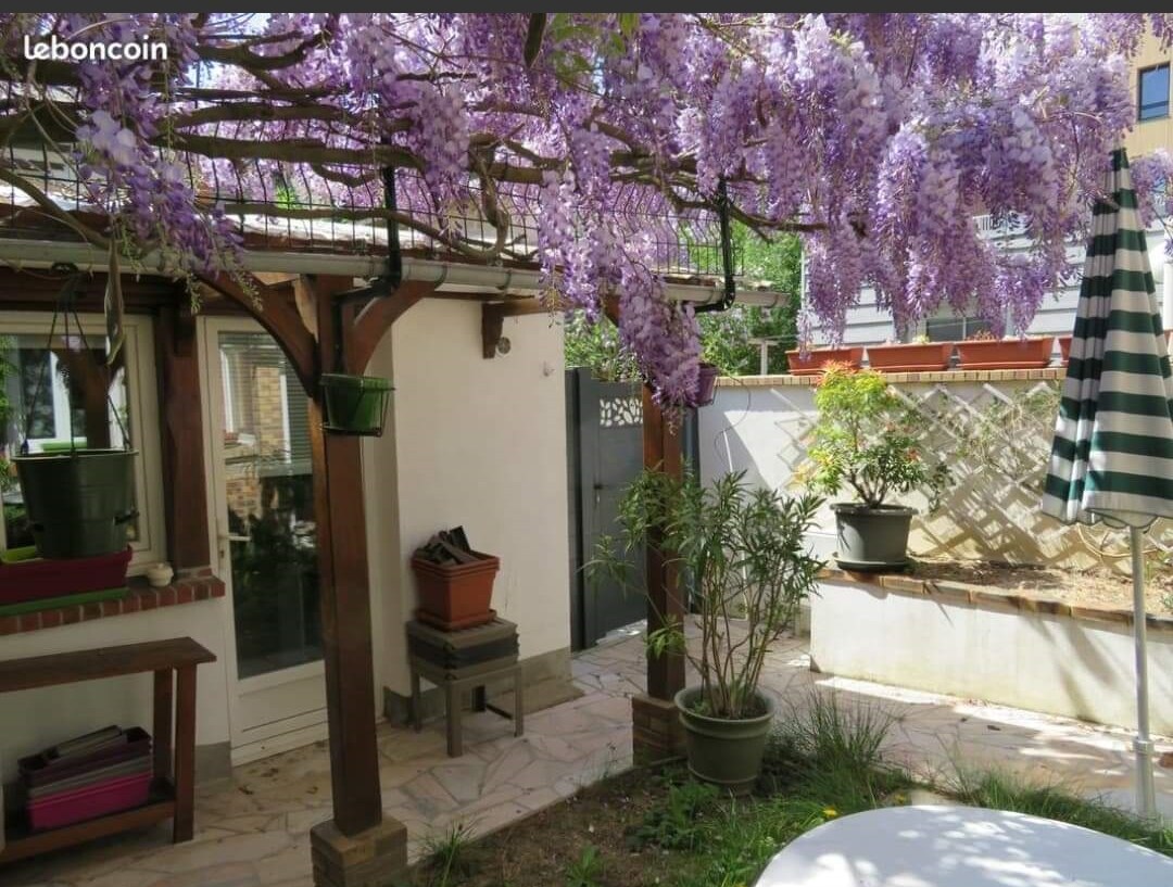 A serene garden space is framed by blooming wisteria, offering vibrant purple clusters above. Potted plants are arranged throughout the area, and a small table with chairs can be seen under one of the pergolas. Natural light filters softly through the leaves.