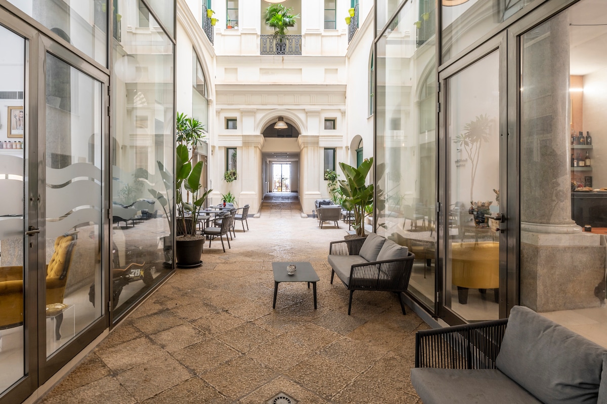 The image captures a courtyard within the Palazzo, featuring large glass doors on either side. Comfortable seating areas with tables are visible, surrounded by potted plants that add greenery. The atmosphere appears calm and serene, with light entering the space, inviting guests to relax.