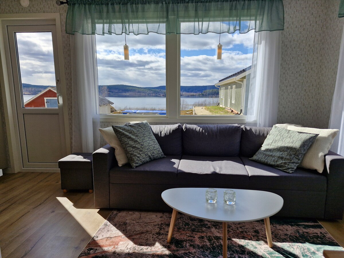 A comfortable living area is highlighted by a dark gray sofa adorned with decorative pillows. A small coffee table sits in front, accompanied by two glasses. Large windows frame a scenic view of the landscape and nearby body of water, bringing natural light into the space.