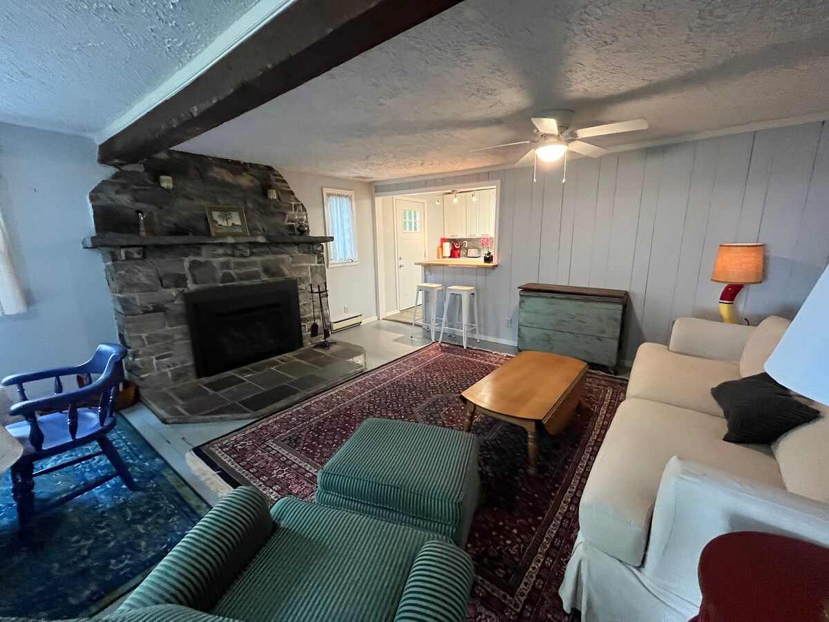 A cozy living room showcases a stone fireplace complemented by a wood beam ceiling. A light-colored sofa is paired with a green striped armchair, and a wooden coffee table sits on an area rug. Natural light enters through a nearby window.