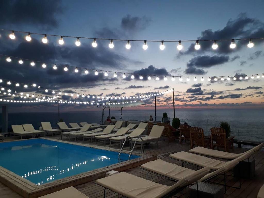 An outdoor area features a swimming pool surrounded by lounge chairs, creating a space for relaxation. String lights hang overhead, illuminating the terrace at dusk. The horizon reveals a blend of colors as the sun sets over the water.