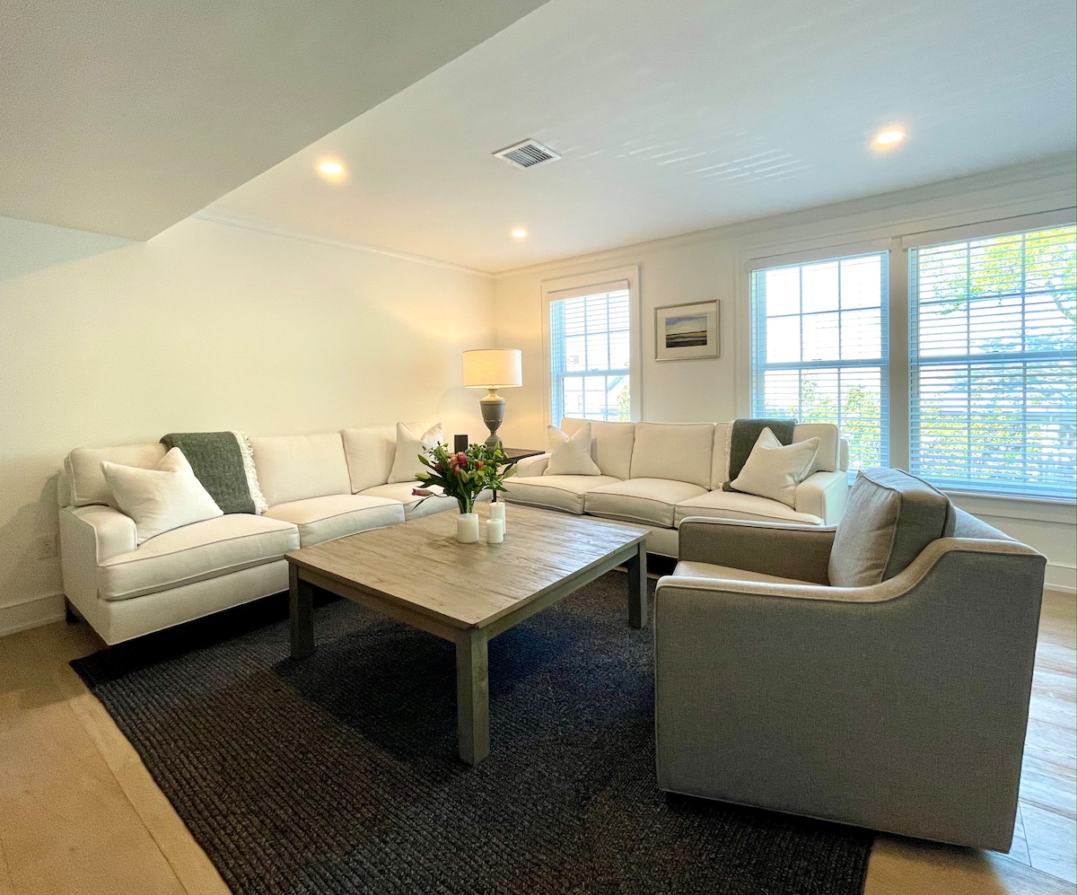 A spacious living area features a large sectional sofa and an armchair, arranged around a wooden coffee table. A light rug anchors the seating arrangement. Natural light enters through multiple windows, while soft lighting from a nearby lamp enhances the space.