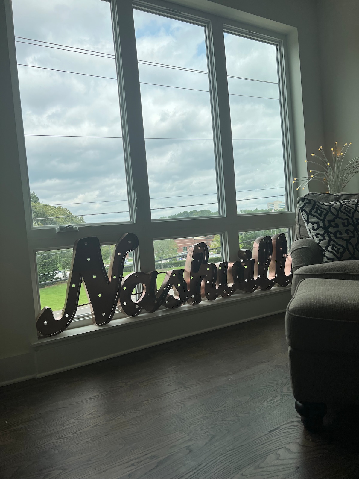 A large, illuminated sign spelling ‘Nashville’ is positioned near a window, casting a warm glow. Natural light filters in through the three-paneled window, highlighting the wooden floor and the plush seating area beside it. Lush greenery is visible outside.