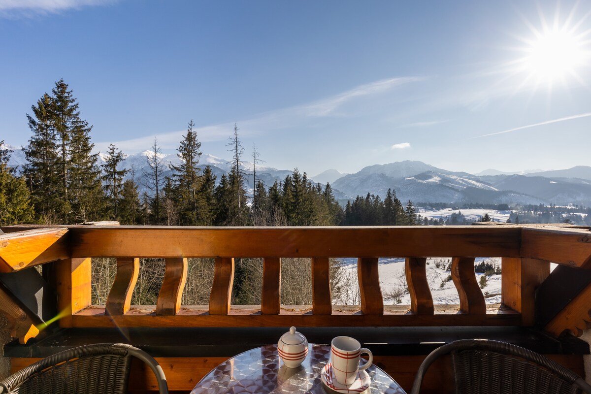 A wooden balcony offers a view of snow-covered mountains and valleys beneath a clear sky. Two cups and a small pot sit on a round table, inviting guests to enjoy the serene surroundings.