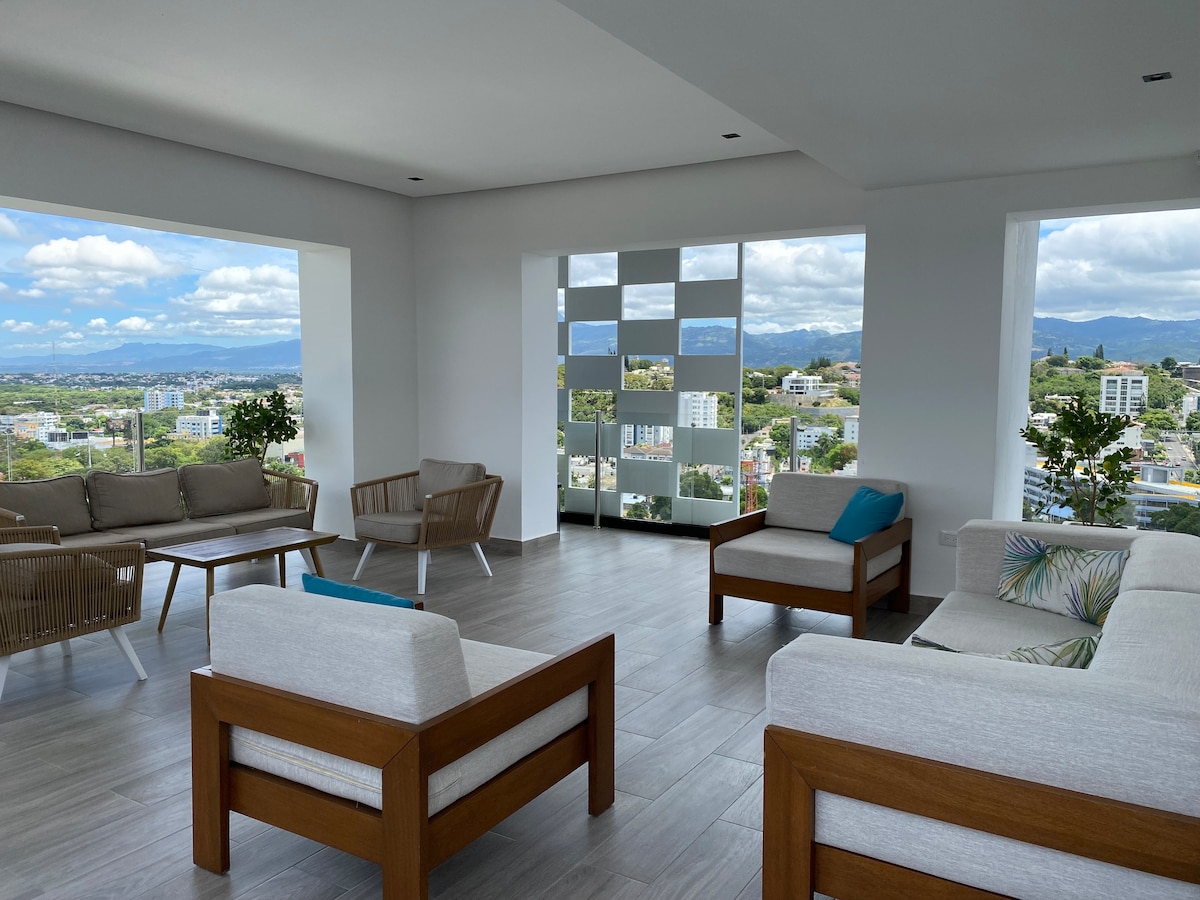 A bright seating area features modern furnishings with light-colored cushions and wooden accents. Large windows provide a view of the surrounding cityscape and greenery. Natural light illuminates the space, enhancing its open and airy feel.
