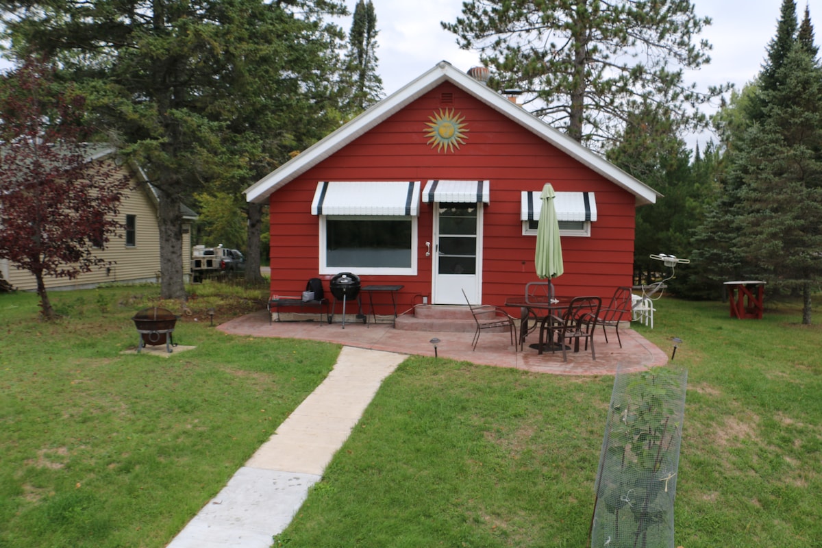 A charming red cottage is surrounded by greenery, featuring a stone patio with outdoor seating. A grill is positioned nearby, and a green umbrella provides shade. The front door is accented by a decorative sun, creating a welcoming entrance.