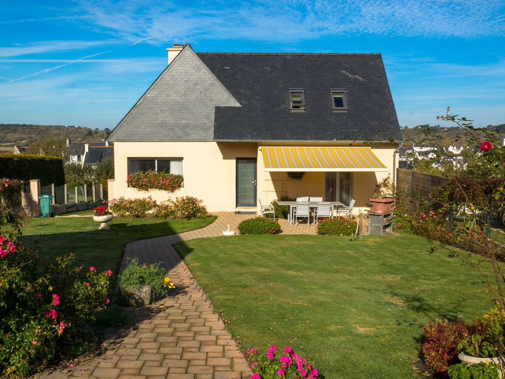A charming independent house is set within a well-maintained garden, featuring a patio area with outdoor seating beneath a yellow awning. Lush green grass and flower beds add color to the outdoor space, while a pathway leads to the entrance of the home.