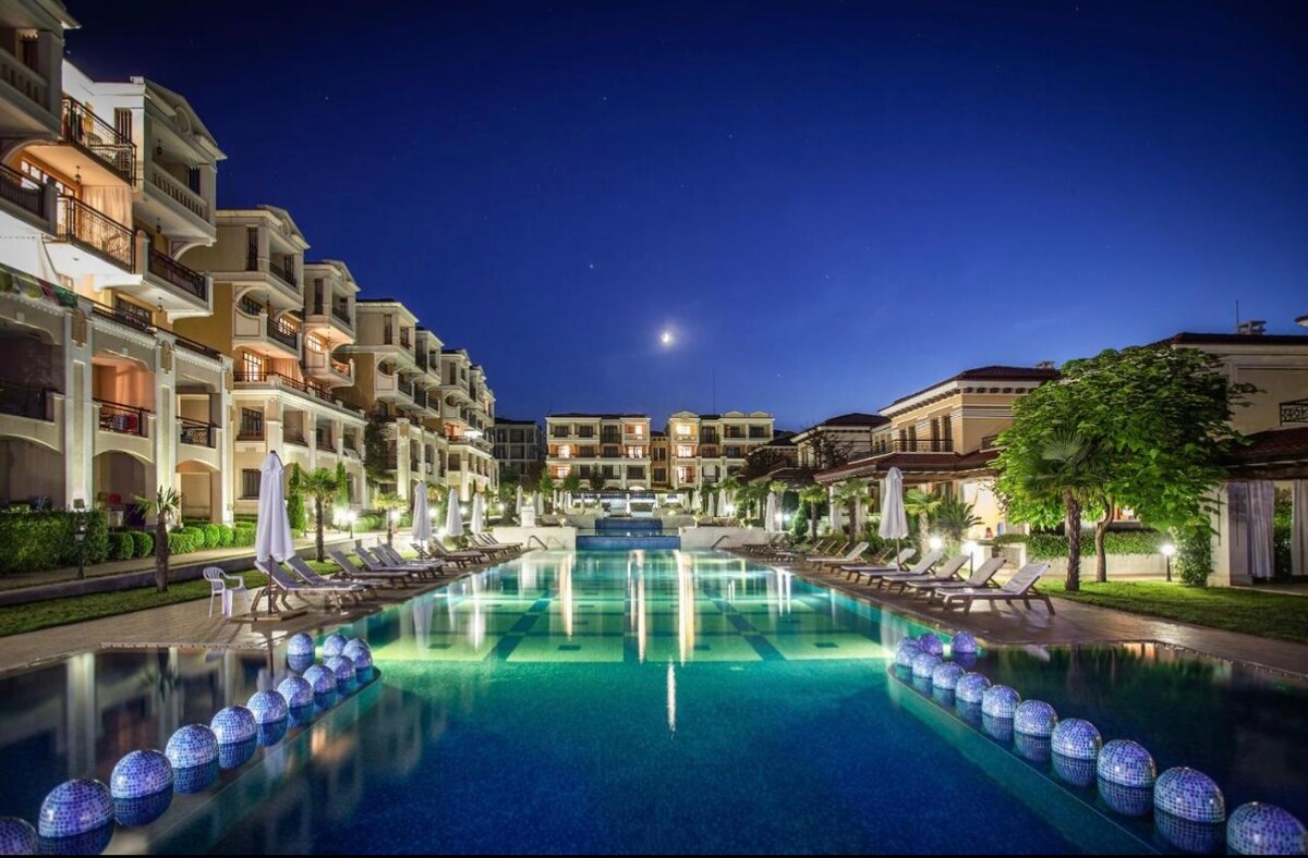 A serene pool area is surrounded by stylish hotel buildings, illuminated under a night sky. Lounge chairs are positioned along the pool, and decorative lights reflect on the water's surface, enhancing the tranquil atmosphere of the resort.