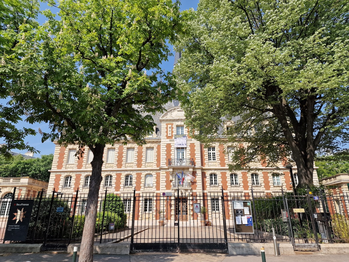The grand facade of a historic building is framed by lush green trees, providing ample shade. A wrought iron gate stands at the entrance, with the building's architectural details accentuated by the sunlight. Flags are displayed prominently, indicating a welcoming atmosphere.
