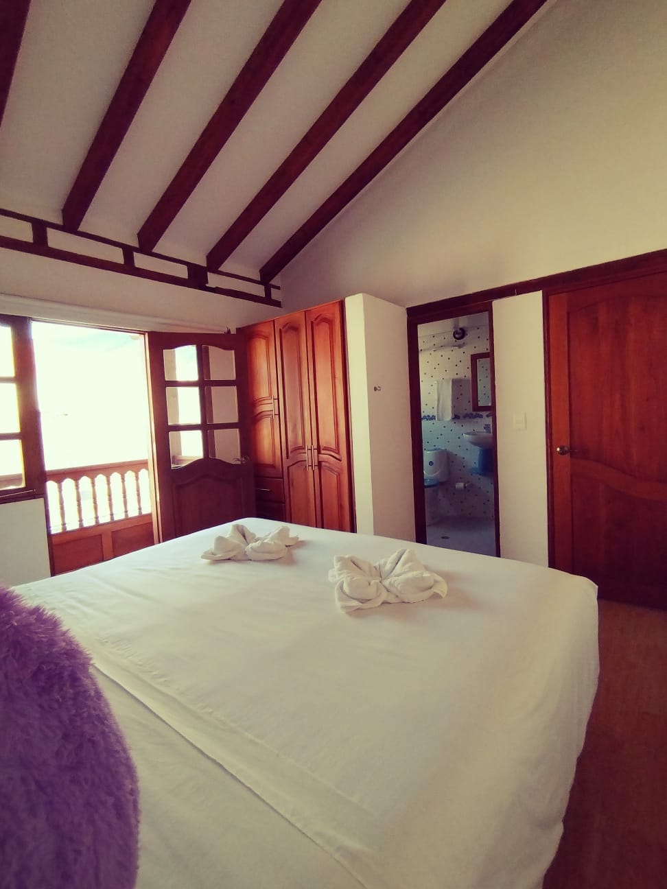 A bright bedroom features a spacious bed adorned with neatly folded towels. Warm wooden accents are visible in the furniture and ceiling beams. Natural light enters through dual windows, with a glimpse of a bathroom reflected in a nearby mirror.