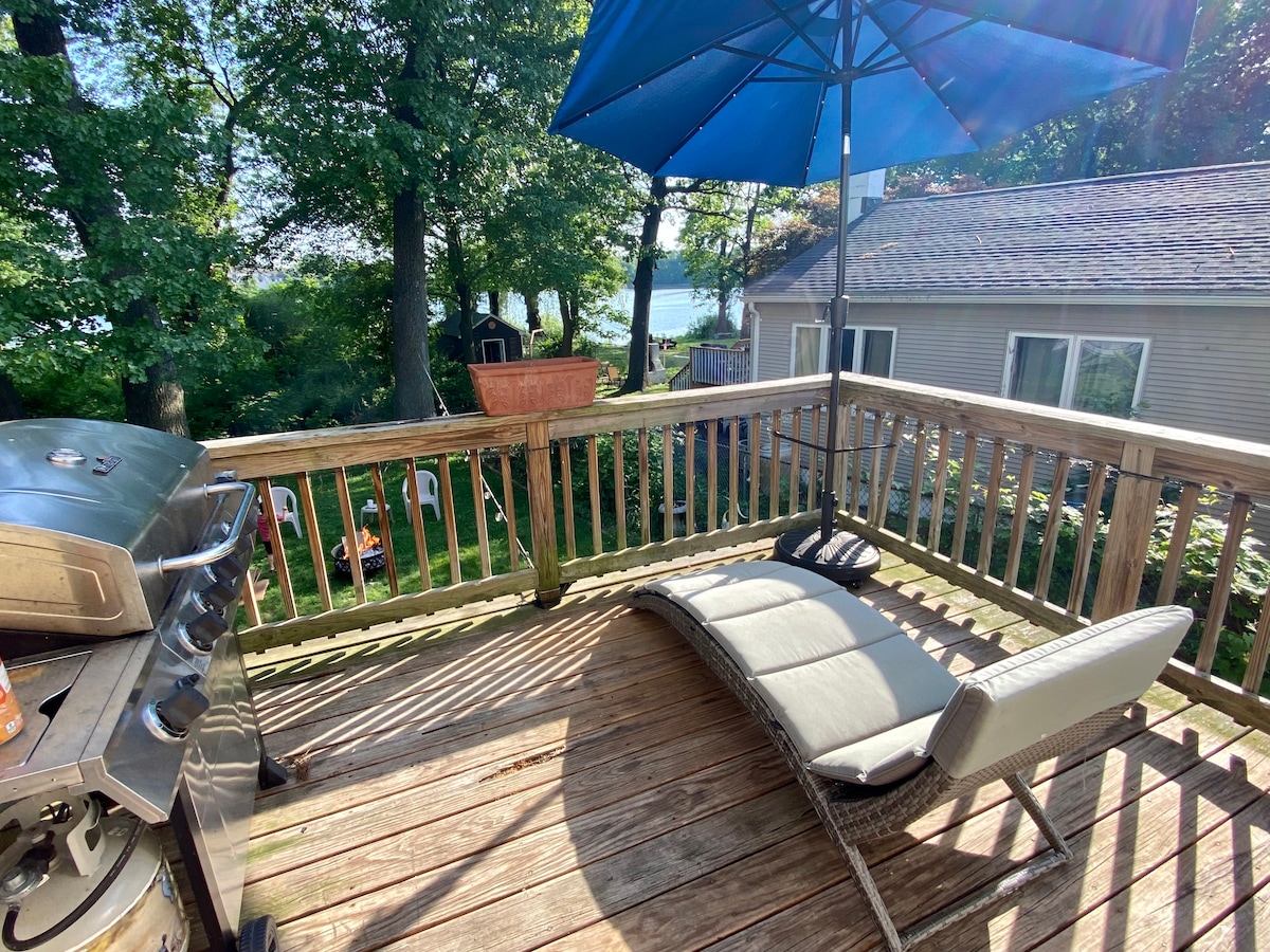A spacious wooden deck is shown, featuring a metallic grill and a lounge chair positioned under a large blue umbrella. Verdant trees provide shade, and views of the pond are visible in the background, creating a serene outdoor setting.