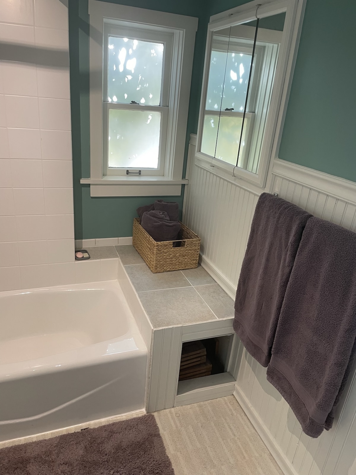 A well-appointed bathroom features a soaking tub alongside a tiled area with a built-in shelf. A basket holds neatly rolled towels, and two plush towels hang from a rack. Natural light filters through frosted window panes, complemented by calming green walls.