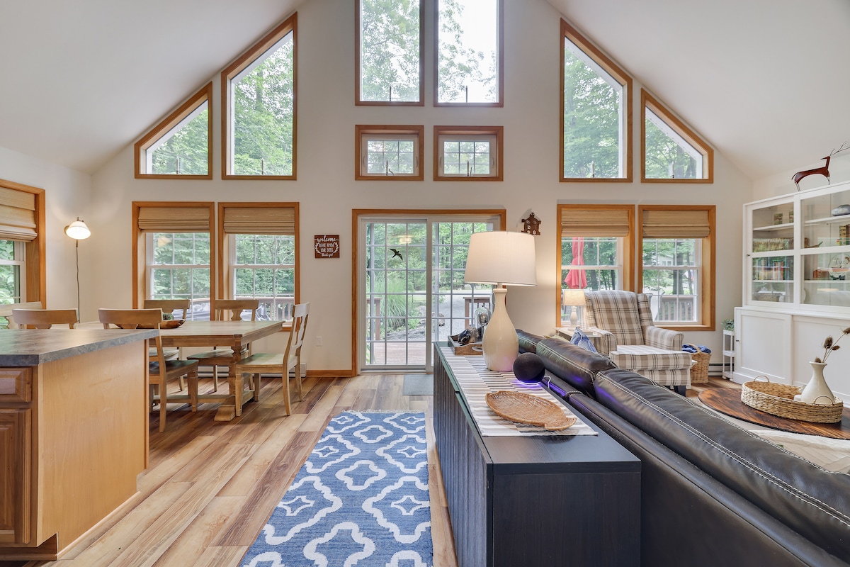 A bright living space features large windows that offer views of the surrounding trees. A cozy sectional sofa is situated near a sleek entertainment center, while a dining area with wooden chairs is visible in the background. Light filters through the spaces, creating a welcoming ambiance.