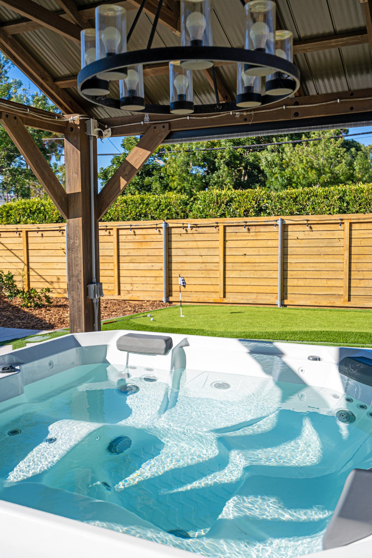 A spacious jacuzzi is featured beneath a wooden structure, complemented by a modern chandelier overhead. The clear, inviting water reflects light, while wooden fencing and lush greenery surround the area, creating a relaxing outdoor space.