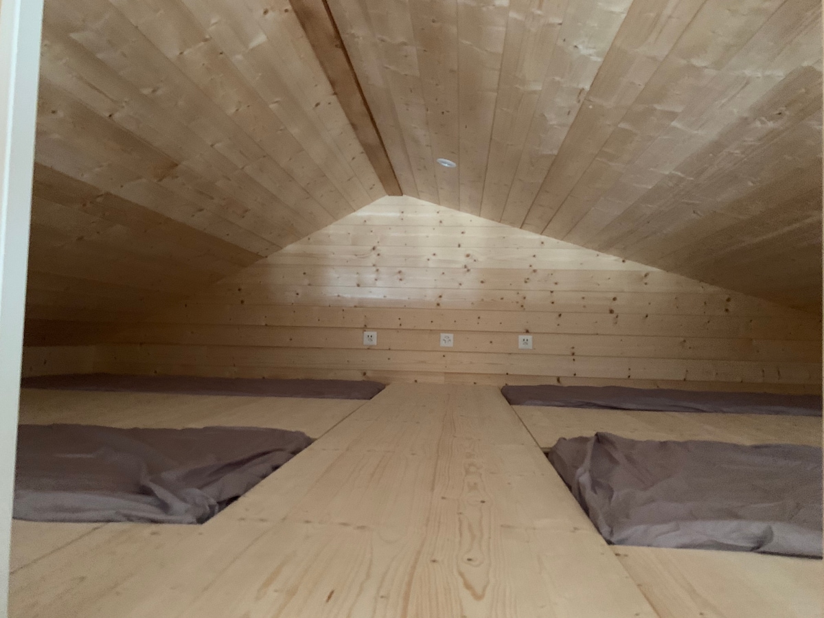 An expansive loft area features wooden walls and a sloped ceiling. Multiple sleeping mats are laid out on the wooden floor, providing an inviting space for rest. Natural light filters through a small opening, softly illuminating the area.