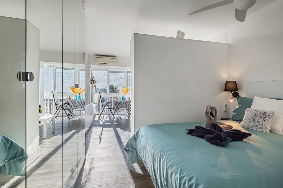 A bedroom featuring a spacious bed with a soothing blue duvet and decorative pillows. Natural light filters in through large windows, illuminating the room. A ceiling fan and a contemporary lamp enhance the modern design. Mirrors reflect the inviting atmosphere of the adjoining terrace.