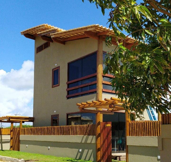 A two-story building features a combination of wooden and stucco materials, highlighted by large windows. A wooden fence surrounds the property, with a well-maintained lawn in front. A clear blue sky complements the structure's earthy tones.