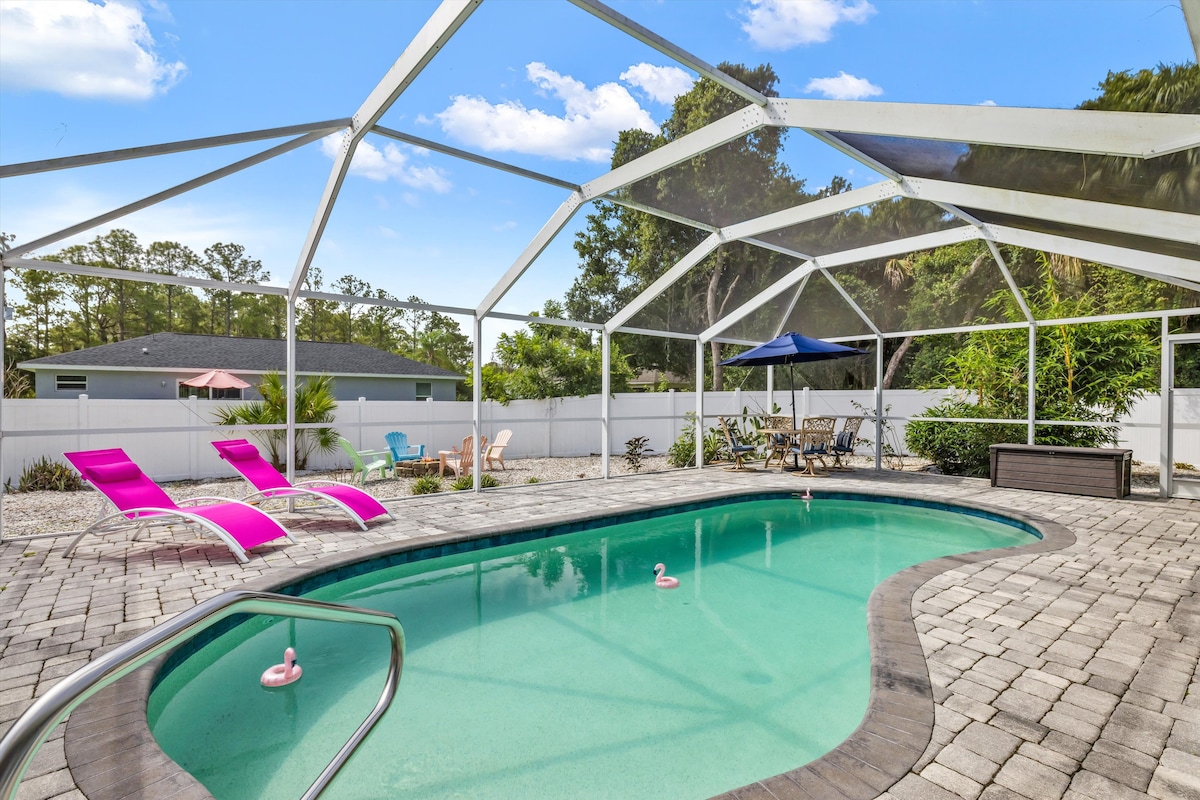 The private saltwater pool is set within an enclosed area, surrounded by a paved patio. Two pink pool floats drift in the water, while two colorful lounge chairs and an outdoor dining table with an umbrella provide options for relaxation and shade.