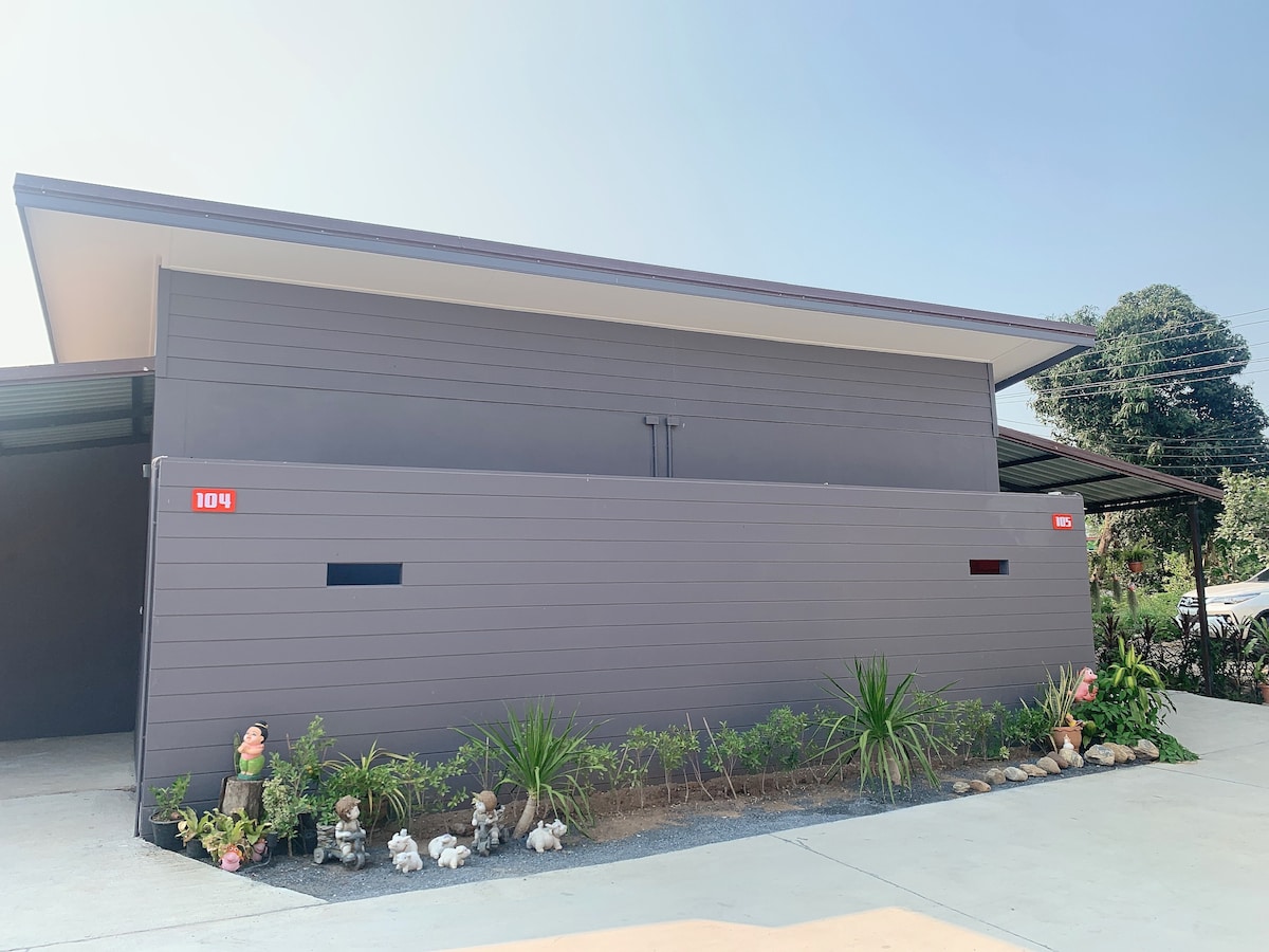 A modern building exterior is shown, featuring a sleek, dark gray facade with minimalistic red signage. Neatly arranged landscaping with various plants and decorative stones enhances the front area, while the clear blue sky above complements the contemporary design.
