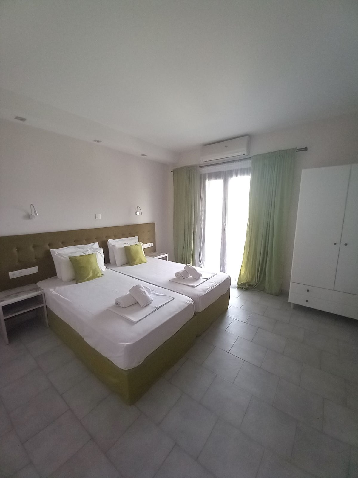 A well-lit room features two spacious beds with crisp white linens and subtle green accents. Light curtains frame the large window, allowing natural light to fill the space. A compact dresser and bedside tables complement the minimalist design, enhancing a sense of openness.