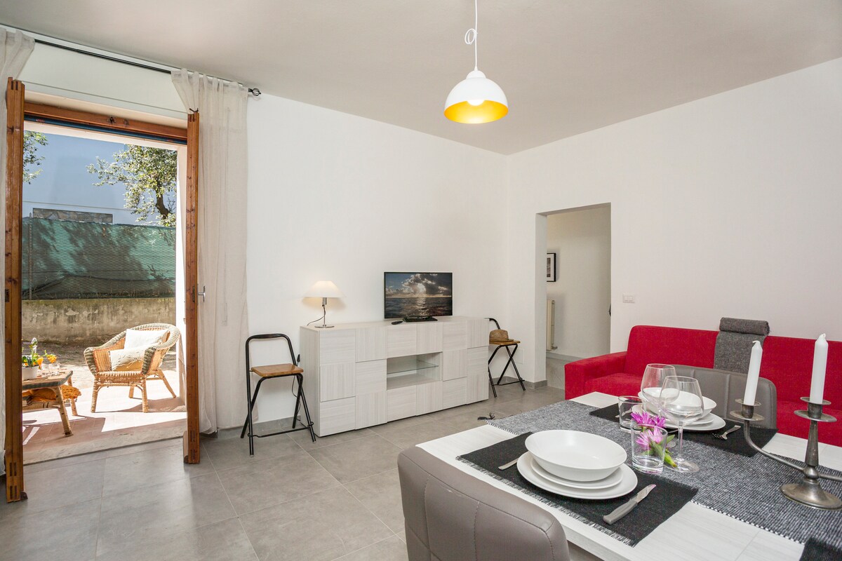 A bright living area features modern furnishings, including a red sofa and a white entertainment unit. A dining table is set with plates and cutlery. Large glass doors open to a private outdoor space, enhancing the connection to the outdoors.