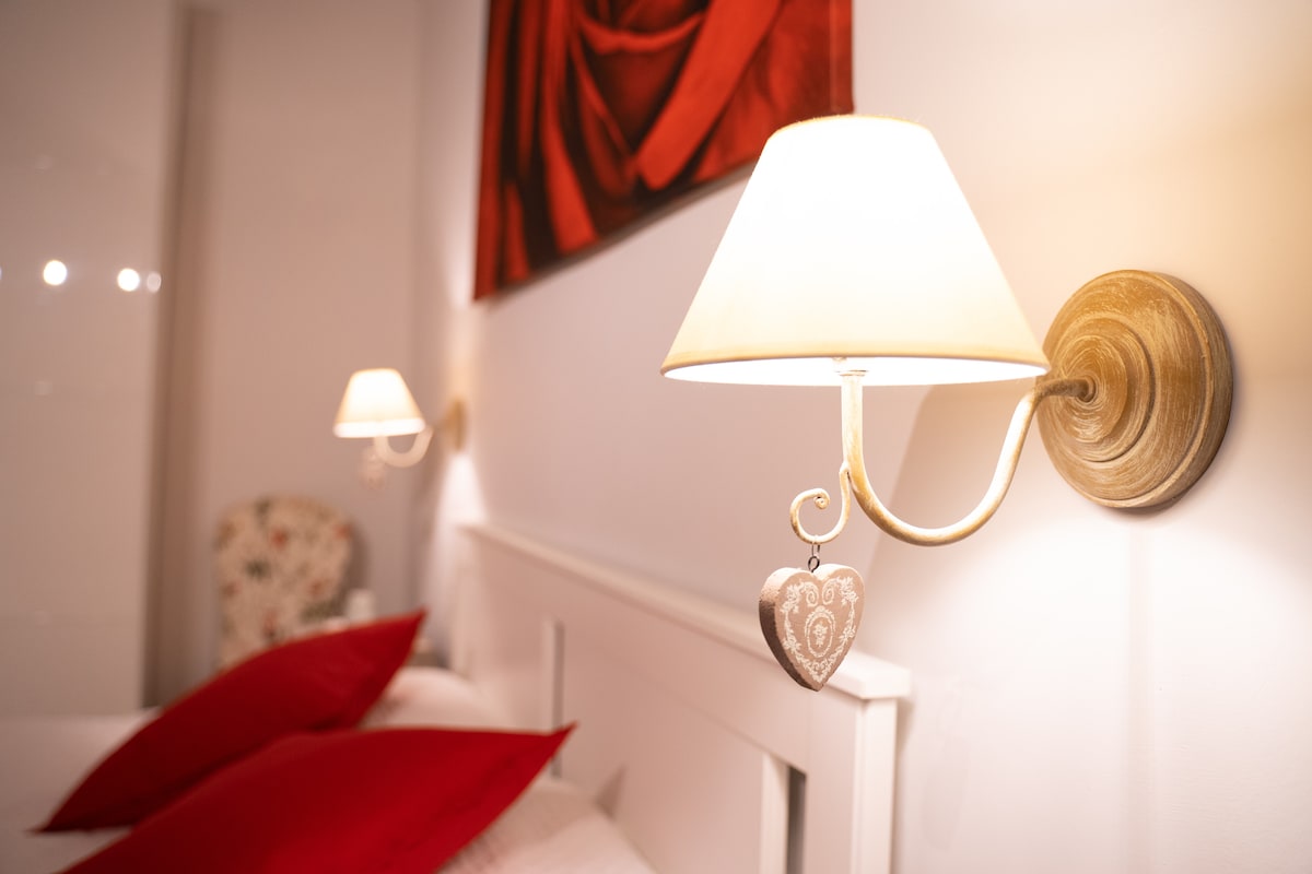 The bedroom features two bedside sconces with soft, warm lighting. The sconces are adorned with decorative heart-shaped elements. Red accent pillows are placed on a spacious bed, while a large piece of wall art depicting a rose adds a subtle touch of elegance.