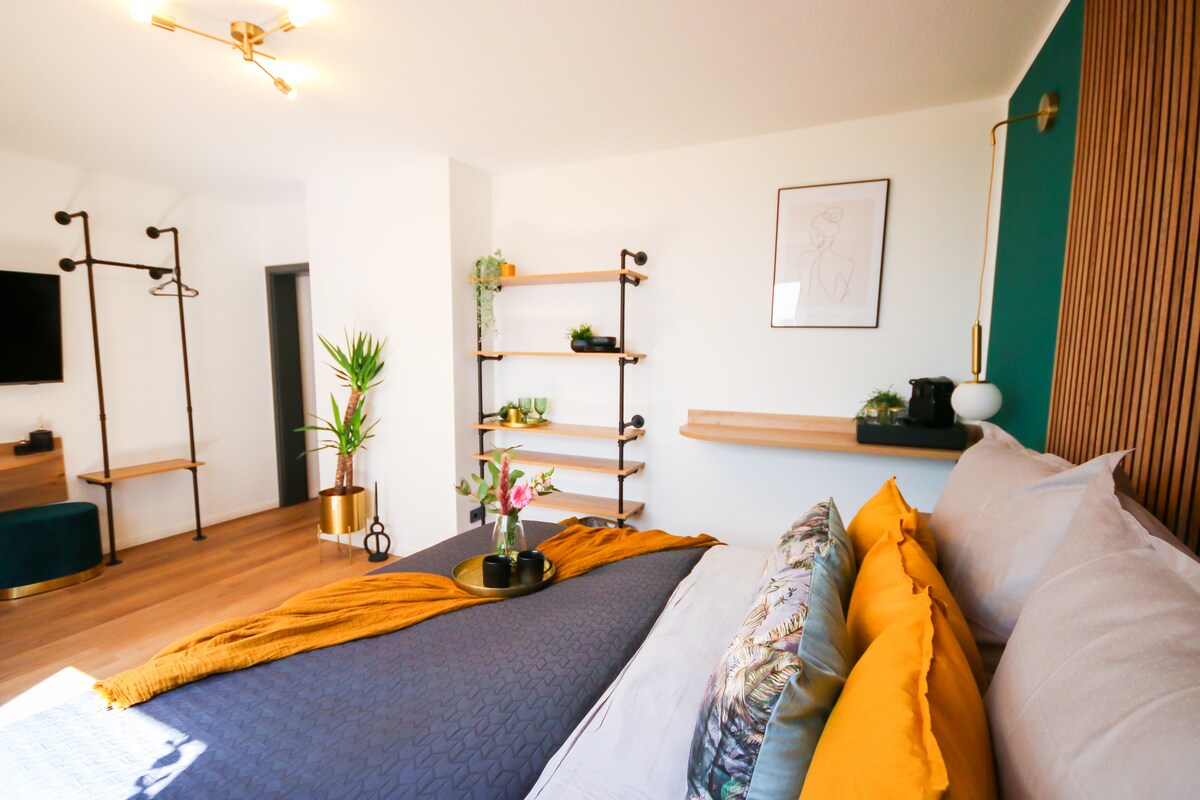 A modern room features a spacious bed adorned with colorful cushions and a textured blanket. Minimalist shelving units and a decorative plant are positioned nearby, while a wall-mounted television and artwork add to the contemporary design.